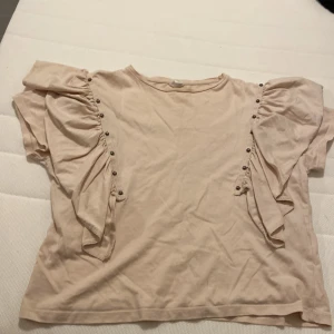 Beige volangblus med nitar från Zara - Supersöt beige blus från Zara med volangdetaljer längs ärmarna och små nitar som ger en cool touch. Blusen har rund halsringning och är tillverkad i mjuk bomull. Perfekt för dig som gillar unika detaljer och en trendig look.