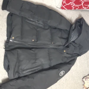 Svart pufferjacka från Canada Goose - Svart pufferjacka från Canada Goose med huva och dragkedja framtill. Jackan har quiltad design, två sidofickor och logotyp på ärmen. Materialet är polyester och dun, perfekt för kalla dagar. Snygg och funktionell stil som håller dig varm. Har haft jackan i 1 år, priset kan diskuteras!