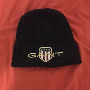 Mörkblå stickad mössa från GANT - Snygg mörkblå stickad mössa från GANT med broderad logga och sköld framtill. Modellen heter Archive Shield Beanie och har en bred uppvikt kant. Perfekt för kalla dagar och ger en cool streetstil. Helt ny lappen finns till och med kvar 