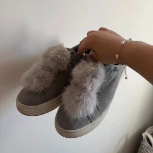 Grå sneakers med fluffig pälsdetalj - Säljer ett par grå sneakers med fluffiga pälsdetaljer på ovansidan. Skorna har platt sula och rund tå, vilket gör dem både snygga och bekväma. Pälsen är ljusgrå och ger en unik och trendig look. Perfekt för dig som vill sticka ut med dina skor.
