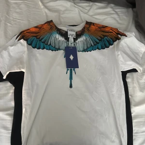 Marcelo Burlon vit t-shirt med vingar - Säljer en vit t-shirt från Marcelo Burlon County of Milan med ikoniskt tryck av färgglada vingar i orange, blått och svart runt halsen och över axlarna. T-shirten har rund hals och korta ärmar, tillverkad i mjuk bomull. Perfekt statement-plagg för dig som gillar streetwear.
