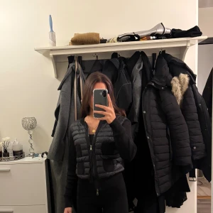 Svart cardigan jacka från Moncler - Svart cardigan jacka från Moncler med quiltad framsida och fyra stickade fickor. Jackan har långa ärmar i stickad bomull, rund halsringning och dragkedja framtill. Klassisk Moncler-logga på ena ärmen. Perfekt för lager-på-lager och snygg till allt.