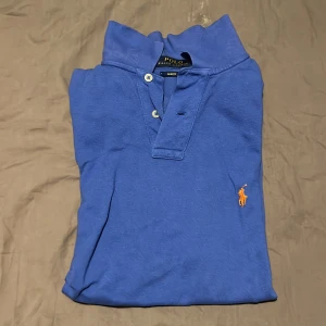 Blå pikétröja Polo Ralph Lauren - Snygg blå pikétröja från Polo Ralph Lauren med klassisk krage, korta ärmar och två knappar framtill. Tröjan har den ikoniska broderade loggan i orange på bröstet och är tillverkad i mjuk bomull. Perfekt för dig som gillar stilrena och sportiga plagg. Strlk M Slim fit