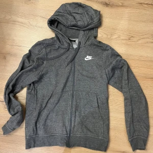 Grå hoodie från Nike med dragkedja - Säljer en grå hoodie från Nike i storlek XS, passar S. Tröjan har huva med snörning, hel dragkedja och två fickor framtill. Nike-loggan i vitt på bröstet ger en clean look. Perfekt för chill eller träning, mjukt material i bomull och polyester.