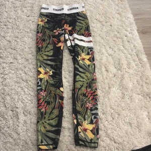 Stronger leggings med blommigt mönster - Snygga leggings från Stronger i storlek XX-Small med tropiskt blommigt mönster i grönt, gult, rött och svart. De har vit elastisk midja med logga och vita ränder på ena benet. Materialet är 85% återvunnen polyester och 15% elastan, perfekt för träning.