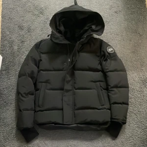 Svart pufferjacka från Canada Goose - Säljer en svart pufferjacka från Canada Goose med huva och klassisk logga på ärmen. Jackan är vadderad och har dragkedja framtill samt ribbade muddar vid ärmsluten. Perfekt för kalla dagar och riktigt snygg streetstil.