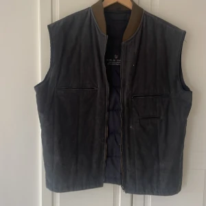 Mörkgrå mockaväst från Gant - Säljer en mörkgrå mockaväst från Gant med quiltad insida och ribbad krage i brunt. Västen har dragkedja framtill och två diskreta fickor med dragkedja. Broderad Gant-logga på ryggen. Perfekt för lager-på-lager-stil.