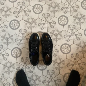 Svarta sneakers från Lanvin med lackad tå - Säljer ett par svarta sneakers från Lanvin med snygg lackad tå och klassisk vit sula. Skorna har snörning och är gjorda i en mix av mocka och skinn, vilket ger en lyxig känsla. Perfekta för dig som vill ha en stilren och trendig look. Låda och dustbag ingår. Fråga om bild om de behövs 
