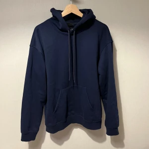Mörkblå hoodie - Säljer en stilren mörkblå hoodie med huva och snörning. Tröjan har en stor magficka och långa ärmar med ribbade muddar. Perfekt för dig som gillar enkel och avslappnad stil. Passformen är normal och materialet känns mjukt och bekvämt.