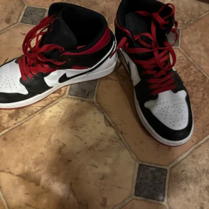 Nike Air Jordan 1 svart/röd sneakers - Säljer ett par Nike Air Jordan 1 sneakers med klassisk high-top design. Skorna har svart, vit och röd färg med röda snören och perforerad tå. Ikonisk Jumpman-logga på plösen och svart swoosh på sidan. Yttermaterialet är skinn och sulan är röd och vit.