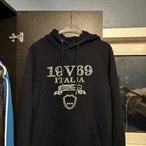 Svart hoodie 19V69 Italia vercage S - Svart hoodie från 19V69 Italia med vit tryckt logga och emblem framtill. Tröjan har huva med snörning och en stor magficka. Perfekt för dig som gillar en stilren och sportig look. Ny pris är 1499kr!!! Har använt den 4 gånger bara säljer då den är för stor för mig 