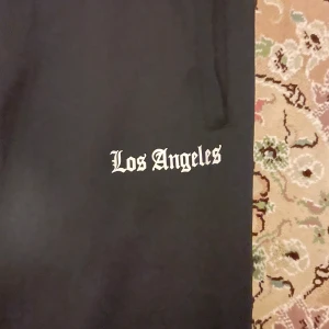 Svarta byxor med Los Angeles-tryck - Svarta byxor med vit sidoremsa och snörning i midjan. På vänster lår står det 'Los Angeles' i vit gotisk stil. Byxorna har en sportig vibe och är perfekta för en chill streetwear-look.