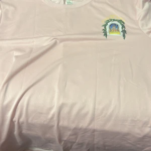 Ljusrosa t-shirt med tryck Casablanca - Säljer en ljusrosa t-shirt med trycket 'Avenida Casablanca' och ett färgglatt motiv av en byggnad i fönsterform med gröna blad runt. T-shirten har rund hals och korta ärmar, tillverkad i mjukt material som känns skönt mot huden.