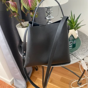 Svart stilren axelväska i skinn från mango - Snygg svart axelväska i minimalistisk design med både axelrem och handtag. Väskan är tillverkad i slätt skinn och har en fyrkantig form som rymmer allt du behöver. Perfekt för dig som gillar en clean och modern look. Väskan är från mango