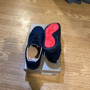 Christian Louboutin marinblå sneakers - Säljer ett par exklusiva marinblå sneakers från Christian Louboutin i mocka med klassisk röd sula och signaturdetaljer undertill. Skorna har snörning, rund tå och platt sula. Kommer med originalkartong och dustbag.