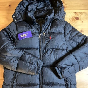 Svart dunjacka från Polo Ralph Lauren - Svart dunjacka från Polo Ralph Lauren med röd broderad logga på bröstet. Jackan har huva, tvåvägsdragkedja och quiltad design. Perfekt för kalla dagar och har en klassisk, sportig look.