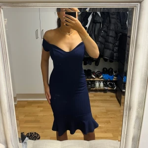 Marinblå offshoulder klänning - Säljer en marinblå klänning med offshoulder-design och v-ringad hals. Klänningen är figurnära med en snygg volangkant nedtill som ger extra stil. Perfekt för dig som gillar en elegant och modern look.
