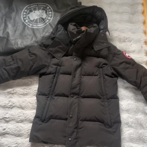 Svart dunjacka från Canada Goose S - Säljer en svart dunjacka från Canada Goose i storlek S. Jackan har luva, klassisk logga på ärmen och är quiltad med stora paneler. Perfekt för kalla dagar och har både dragkedja och knappar framtill. Snygg och tidlös design som håller värmen. Pris kan diskuteras och vid flera bilder sa ba att höra av sig.