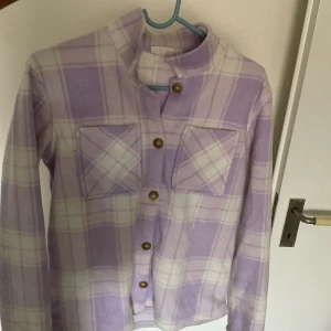 Ljuslila rutig skjorta från KappAhl - Supermysig skjorta från KappAhl i ljuslila och vitt rutigt mönster. Den har två stora bröstfickor, bruna knappar framtill och är långärmad. Materialet är mjukt och fleece-liknande, perfekt för chill dagar. Snygg ståkrage för extra stilpoäng. ( Jag säljer den då den inte är min stil längre )
