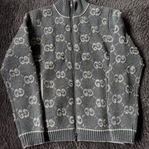 Grå kofta med GG-mönster från Gucci - Snygg grå kofta från Gucci med hel dragkedja och ribbad krage. Tröjan har ett ikoniskt GG-mönster i vitt över hela plagget och ribbade muddar vid ärmslut och nederkant. Perfekt för dig som gillar lyxiga detaljer och klassisk design.