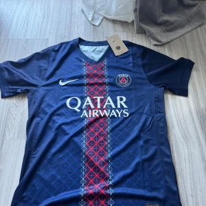 PSG Hemmaställ 25/26  - Paris Saint-Germain fotbollströja från Nike i marinblått med rött och vitt geometriskt mönster längs mitten. Kortärmad med rund hals och PSG-logga på bröstet. Materialet är lätt och andas, perfekt för träning eller match. Qatar Airways tryck på framsidan.