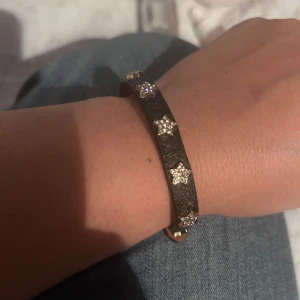 Svart armband med glittriga stjärnor - Snyggt svart armband med små glittriga stjärnor i guldfärg längs hela bandet. Armbandet har ett smalt läderliknande band och ett metallspänne i guld. Perfekt för dig som gillar detaljer med lite extra bling och stjärnformade dekorationer.
