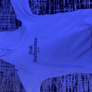 Vit hoodie Peak Performance - Snygg vit hoodie från Peak Performance med svart tryck på bröstet. Klassisk huva och stor magficka framtill. Mjuk insida i bomullsmix, perfekt för chill eller häng. Stilren och enkel design som funkar till det mesta.