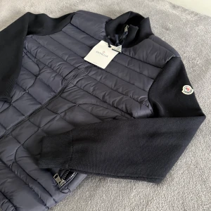 Moncler cardigan  - Snygg marinblå cardigan från Moncler med quiltad front och stickade ärmar. Jackan har dragkedja, ribbad krage och muddar samt Moncler-logga på ärmen. Materialmix  och stickat ger en modern vibe. Perfekt för dig som vill ha en clean och stilren look.
