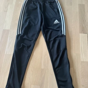 Svarta Adidas träningsbyxor med dragkedja - Svarta träningsbyxor från Adidas med klassiska vita ränder längs sidorna och logga på låret. Byxorna har smal passform, elastisk midja och dragkedja vid bensluten. Tillverkade i polyester med Climacool-teknologi för extra komfort.