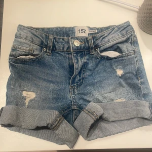 Blå jeansshorts från 157, stl 140 - Snygga blå jeansshorts från 157 med upprullade benslut och slitna detaljer framtill. Klassisk femficksmodell och knappgylf. Perfekta för varma dagar och enkel att matcha med allt. Märke: 157, modell Sunny Star.