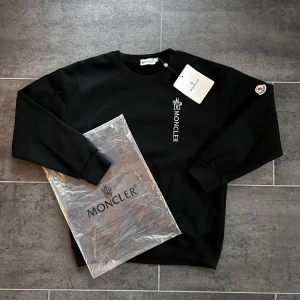 Svart tröja från Moncler - Svart sweatshirt från Moncler med vit broderad logga på bröstet och klassisk Moncler-patch på ärmen. Tröjan har rund hals, långa ärmar och ribbade muddar. Materialet är mjuk bomull och passformen är normal. 