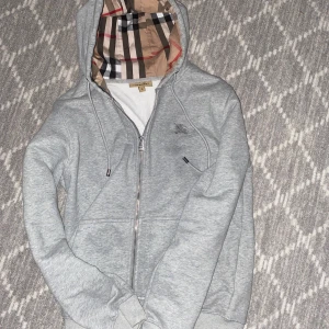 Burberry zip-up hoodie  - Grå hoodie från Burberry i bomull med klassiskt rutigt foder i huvan. Tröjan har dragkedja framtill, två fickor och broderad logga på bröstet. Perfekt för dig som gillar stilren design med en touch av lyx.