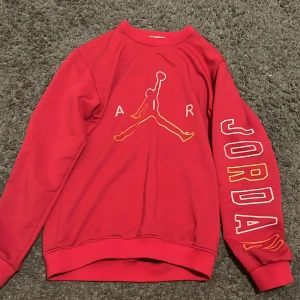 Röd Jordan sweatshirt med tryck - Säljer en röd Jordan sweatshirt med stort Air Jordan-tryck på bröstet och texten 'JORDAN' längs ena ärmen i vitt och gult. Tröjan har rund hals, långa ärmar och ribbade muddar. Perfekt för dig som gillar sportig streetstyle.