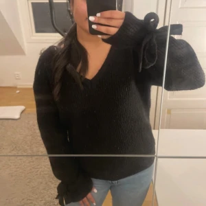 Svart stickad tröja med knyt från NA-KD - Mysig svart stickad tröja med v-ringning från Linn Ahlborg x NA-KD. Tröjan har långa ärmar med snygga knytband vid ärmsluten som ger en extra touch. Perfekt för dig som gillar stilrena detaljer och vill ha något som känns både chill och trendigt.