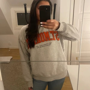 Grå sweatshirt  - Säljer en grå sweatshirt med trycket 'ST. MORITZ' i orange och svart på bröstet. Tröjan har rund hals, långa ärmar och ribbade muddar vid ärmslut och nederkant. Mjuk och skön, perfekt för chill dagar. Materialet är bomull och polyester.