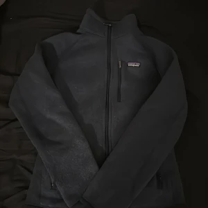 Mörkblå fleecejacka från Patagonia - Mysig mörkblå fleecejacka från Patagonia med hög krage och hel dragkedja framtill. Jackan har en bröstficka med dragkedja och två sidofickor. Perfekt för kyliga dagar och enkel att matcha med det mesta.