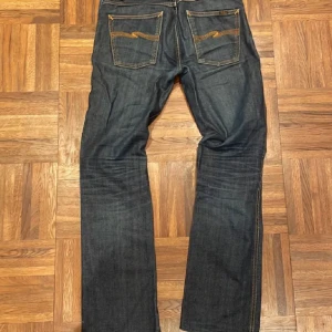 Nudie Jeans blå denim - Mörkblå jeans från Nudie Jeans med klassisk orange söm och broderad bakficka. Raka ben och normal passform, tillverkade i slitstark bomullsdenim. Perfekta för dig som gillar stilrena och tidlösa jeans.