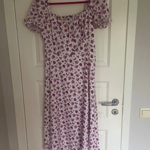 Vit långklänning med lila blommor - Supersöt långklänning i vitt med lila blommigt mönster och puffärmar. Klänningen har snörning vid bysten och en luftig passform, perfekt för varma dagar💌