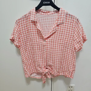 Rutig rosa kortärmad skjorta med knyt - Kortärmad skjorta i rosa och vitt rutigt mönster. Skjortan har klassisk krage, knappar framtill och en snygg knytning nedtill för en chill vibe. Perfekt till sommaren och lätt att matcha med jeans eller shorts.