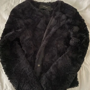 Svart pälsjacka från Vero Moda - Mjuk och fluffig svart pälsjacka från Vero Moda med rund hals och diskreta knappar framtill. Jackan har lång ärm och är perfekt för dig som vill ha en cool och mysig look under kyliga dagar.