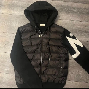 Moncler Cardigan - Säljer en svart cardigan jacka från Moncler med vadderad framsida och huva. Jackan har dragkedja två fickor fram och en vit detalj på ena armen. Jackan har NFC på loggan och tag som man kan scanna. Inte använd. Passar som S-M