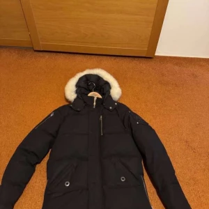 Svart dunjacka med pälsluva Moose Knuckles - Säljer en svart dunjacka från Moose Knuckles med fluffig vit päls runt luvan. Jackan har flera dragkedjor, knappar och fickor samt en snygg patch på ärmen. Perfekt för kalla vinterdagar och har en riktigt schysst puffer-look. Äkta