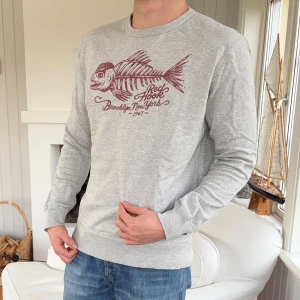 Grå sweatshirt från Ralph Lauren denim & supply - Grå sweatshirt med tryck av fiskben och texten 'Red Hook Brooklyn, New York 1967' i vinrött på bröstet. Rund halsringning och långa ärmar. Tillverkad i 100% bomull, perfekt för chill dagar och enkel att matcha med jeans.