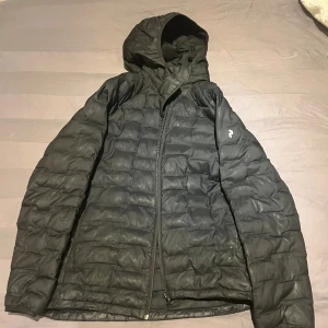 Svart dunjacka Peak Performance 170 - Svart dunjacka från Peak Performance med huva och quiltad design. Jackan har dragkedja framtill och är långärmad, perfekt för kalla dagar. Stilren look och lätt att matcha med olika outfits. Passar dig som gillar funktion och enkel stil.