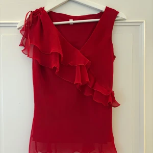 Röd volangblus  - Säljer en snygg röd blus med v-ringning och dubbla volanger framtill. Blusen är ärmlös och har ett omlottliknande fall med transparenta detaljer i chiffong.  Säljer då den är för liten för mig. 