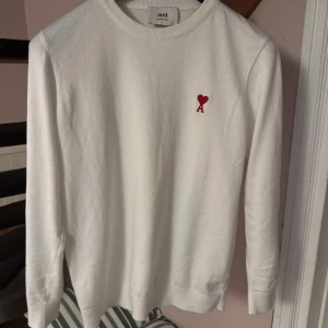 Vit stickad tröja från Ami - Vit stickad tröja från Ami med rund hals och lång ärm. På bröstet finns ett rött broderat hjärta med bokstaven A. Tröjan har en clean och stilren look, perfekt för dig som gillar minimalistisk design och snygga detaljer.
