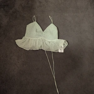 Mintgrön linnetopp med volang - Superfin mintgrön linnetopp med smala axelband, knyt i ryggen och volangkant nedtill. Toppen har ett diskret prickigt mönster och är gjord i lätt polyester.