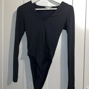 Svart långärmad bodysuit med v-ringning - Säljer en svart långärmad bodysuit med snygg v-ringning. Perfekt att styla med jeans eller kjol för en clean look. Bodyn har knäppning nedtill och är gjord i mjukt stretchigt material som sitter tight mot kroppen.