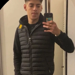 Svart dunväst med gul detalj - Säljer en svart dunväst från Parajumpers med horisontella quiltade paneler och en gul rem vid axeln. Västen har dragkedja framtill och en stilren, sportig look som passar perfekt till hoodie eller t-shirt.