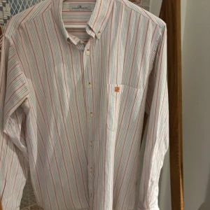 Randig skjorta från Carolina Herrera - Snygg skjorta från Carolina Herrera i ljusrosa med tunna ränder i blått, grönt och vitt. Klassisk krage, knappar framtill och broderad CH HC-logga på bröstfickan. Skjortan är långärmad och har en stilren, normal passform. Perfekt för dig som gillar preppy och färg. OBS, saknar nedersta knappen men reser knapparna man kan sy på medföljer.